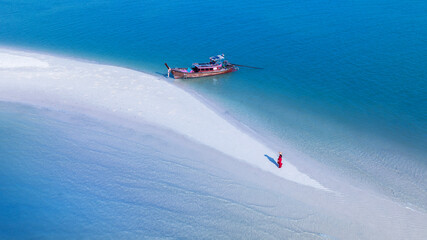A girl in a red dress walking on a white beach with a long-tailed boat moored on the beach with...