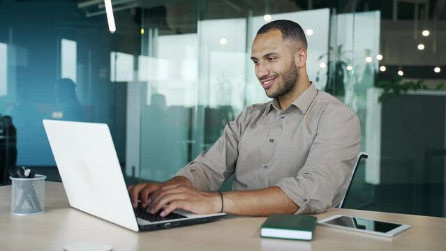 A young smiling businessman is typing on a laptop while sitting at a workplace at a desk in a modern office. Handsome mixed race male working on a project. Employee corresponds with the client online