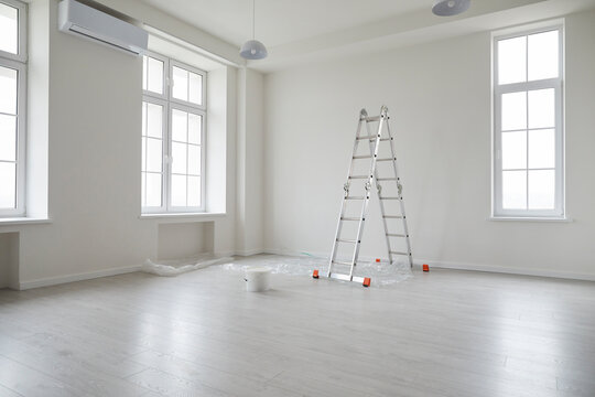 Modern apartment after renovations. New empty office interior or living room at home with white freshly painted walls, step ladder, paint bucket, plastic on the floor, big windows, and air conditioner