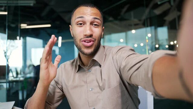 POV of handsome worker making selfie video call while sitting in modern office. The man looks at the camera of the smartphone and cheerfully waves his hand. Businessman is talking online with a client