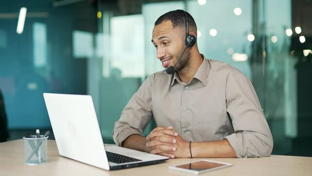 A corporate operator who works in customer support speaks on a video call using a laptop. Call center agent in wireless headset helping customer with complaints in modern office. Online consultation