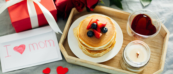 Banner image for desing, web page. Mother's Day concept. Pancakes with berry, tea cup, burning candle, flowers and gift box. Top view