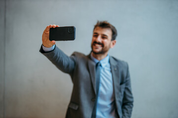 Smiling suited businessman standing against the gray background and taking a selfie.