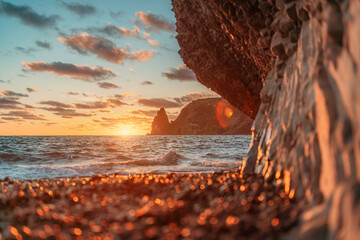Sea sunset waves with foam. Waves run on pebbles and rock from volcanic basalt. Water breaks into splashes and white foam. Against the background of the cape and the sunset sky.