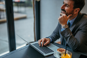 Shot of a young smiling businessman using a laptop, looking confident, and having coffee in a modern workplace.