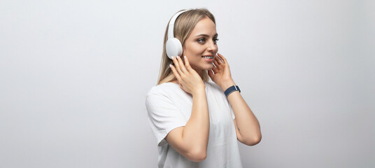 horizontal photo of a young woman in a white tank top listening to a podcast in wireless headphones on a white background with free space