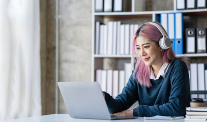 Beautiful Asian businesswoman wearing headphones talking via video call on Her notebook and smartphone to record product information, income, and online business idea.