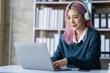 Beautiful Asian businesswoman wearing headphones talking via video call on Her notebook and smartphone to record product information, income, and online business idea.