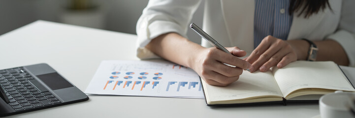Confident Asian businesswoman sitting and taking notes in financial book Income tax with laptop computer or tablet in a happy office.
