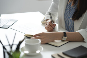 Confident Asian businesswoman sitting and taking notes in financial book Income tax with laptop computer or tablet in a happy office.