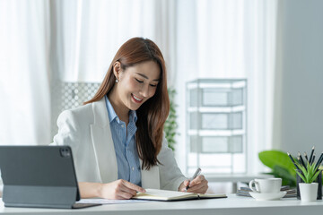 Confident Asian businesswoman sitting and taking notes in financial book Income tax with laptop computer or tablet in a happy office.
