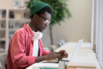 Young African American man freelancer using public library as workspace, typing on laptop. Focused...