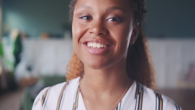 Young happy beautiful African American woman millennial with curly hair looks at camera with smile and turns head little demonstrating good mood dressed in casual style stands in room. Close up