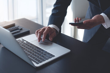 Businessman working on laptop computer at modern office on table, closeup. Business man using mobile phone and typing on laptop, searching the information, internet networking, corporate business