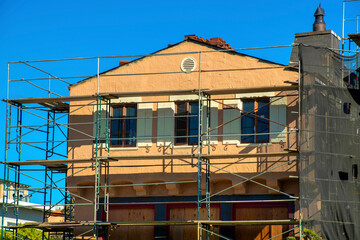 Adobe style house design with shutters and double gable roof under construction with metal scafolding in sun blue sky