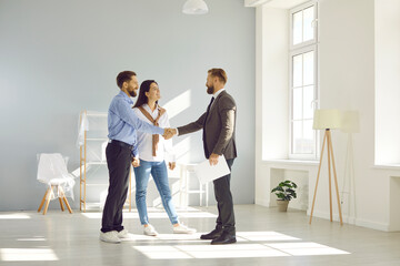 Young married couple make deal, buy new house and exchange handshake with relator. Happy man and woman standing in modern light spacious light living room and shaking hands with real estate agent - obrazy, fototapety, plakaty