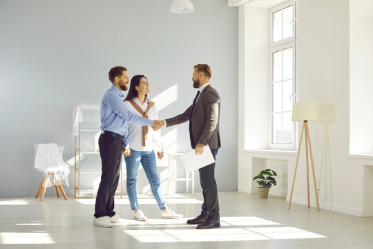 Young married couple make deal, buy new house and exchange handshake with relator. Happy man and woman standing in modern light spacious light living room and shaking hands with real estate agent