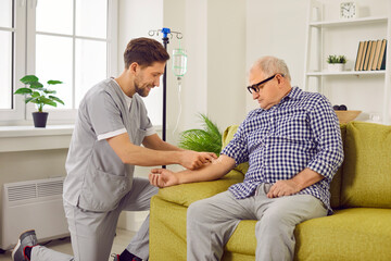 Doctor gives intravenous infusion to senior patient. Young nurse in scrubs uniform inserts IV line needle in vein of old man sitting on sofa at home. Medicine, vitamin therapy, medication concept - obrazy, fototapety, plakaty