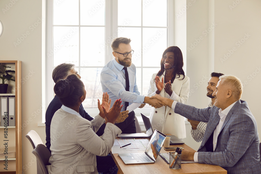 Wall mural Two men shaking hands celebrating success, making a deal, business achievement or signing a contract on their workplace. Group of business people in office applauding to their coworkers.