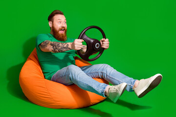 Full length photo of impressed funky guy dressed t-shirt sitting bean bag driving car empty space isolated green color background