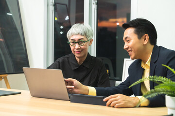 Senior businesswoman and middle aged businessman professionals working late overtime together at night in office. Business people meeting with laptop and tablet computer with happy face in workplace