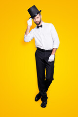 Full size fullbody portrait of stylish attractive magician in tophat, formal wear, looking at camera, staying leg by foot, holding hat with hand, isolated on grey background