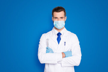 Portrait of attractive handsome dentist with hairstyle in protective face mask, white lab coat, blue tie, holding equipments in crossed arms, isolated on grey background