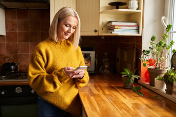 Smiling mature woman using phone texting message while standing in a kitchen
