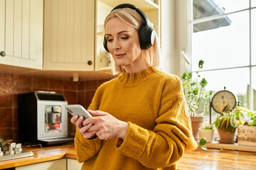Adult woman using phone app listening  music on wireless headphones in a kitchen
