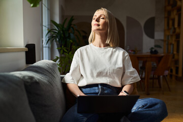 Mindfulness concept. Worek life balance. Adult woman with laptop relaxing on the sofa with closed eyes - obrazy, fototapety, plakaty