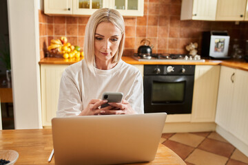 Mature woman doing online payments with phone and laptop sitting at table in a kitchen