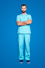 Fototapeta premium Full size body portrait of attractive concentrated man in blue lab suit, having his arms crossed, looking at camera, isolated on grey background