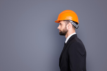 Profile side view half face portrait with copy space, empty place of virile stylish investor with stubble in orange safety helmet, hardhat and black tux with tie, isolated on grey background