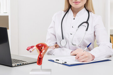 Young beautiful doctor woman working happy and smile in hospital, sitting on table. Model of female reproductive system on work desk. - obrazy, fototapety, plakaty