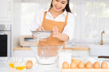 Young woman wearing light brown apron in the modern kitchen have fun sifting bread flour, preparing different ingredients before mixing, and kneading before baking in oven to make homemade bread.