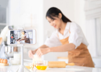 Obraz na płótnie Canvas Portrait of concentrated young woman food vlogger showing process of baking in live stream, standing in front of smart phone on tripod, broadcasting. Indoor studio shot on kitchen background.
