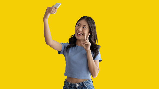 Cheerful young woman taking a selfie with a mobile phone and raising two fingers, Portrait of happy girl with bright face isolated on yellow background, Take a photo.