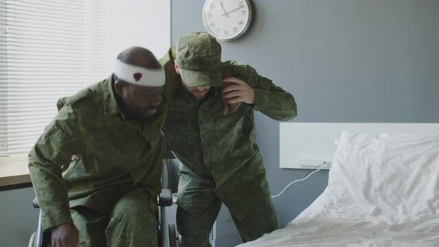 Caucasian lieutenant helping his injured Black friend to get to bed in military hospital room