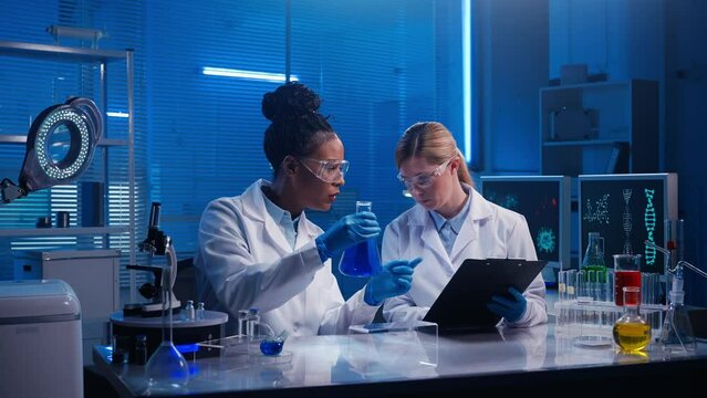 An African American female doctor examines a blue liquid in a flask while a white woman laboratory assistant writes down the data. Medical workers in white gowns work in a lab with blue light.