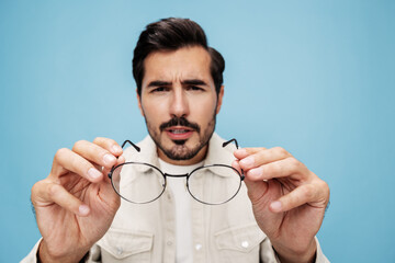 Fototapeta premium Close-up portrait of a brunette man looking through glasses that he holds in his hands, eye problems, glasses for vision farsightedness and nearsightedness, on a blue background, copy space 