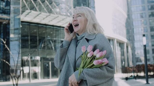 A senior woman talking by phone holding tulips outdoors