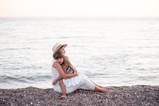 Little girl sits in the arms of pregnant mother on the sand by the sea on beach. Daughter hugging millennial woman in hat. The journey of single parent. Travel pregnancy, copy space. Summer vibes