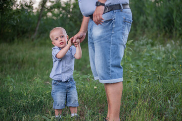 Portrait of little son holding his fathers hand, looking at the camera. Man and boy are dressed in blue jeans. Walk through the forest in the countryside. Care of the child. Children Protection Day