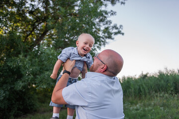 Bald man with glasses throws child into the sky air. Father in jeans plays, embrace with son in nature outside the city. Little boy laughs, having fun with dad on journey in the forest. Fathers day