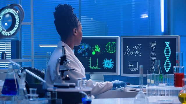 A young African American woman types on a computer keyboard and then examines the sample under a microscope. A black female doctor works in a modern biochemical laboratory with blue light.