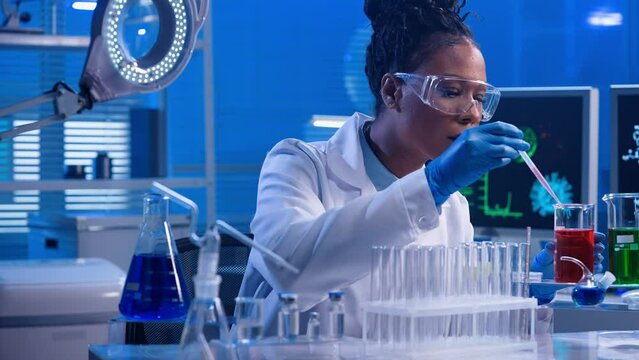 A young African American woman using a plastic pipette pipettes red and green liquid into glass test tubes. A black female doctor works in a modern biochemical laboratory with blue light.