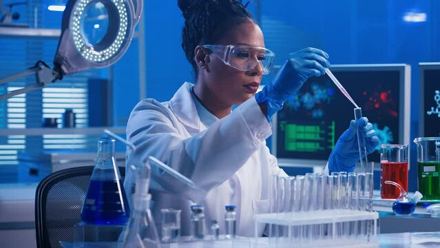 A young African American woman using a plastic pipette pipettes red and green liquid into glass test tubes and examines samples. A black female doctor works in a biochemical laboratory with blue light