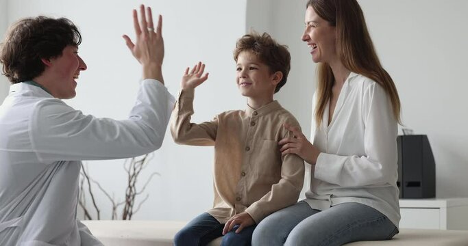 Happy pediatrician man in white coat meeting with cheerful cute little patient boy and mom for examination in office. Male doctor talking to child and parent, laughing, giving high five