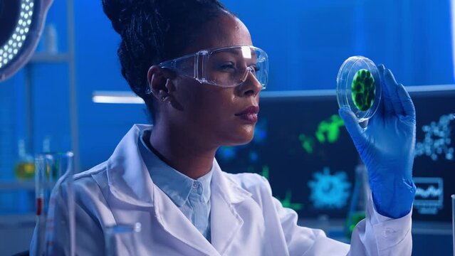 A young African American woman examines a petri dish with a green sample of cultured bacteria. A black female doctor works in a modern biochemical laboratory with blue light. Close up.