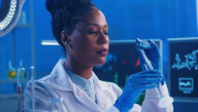 A young African American woman pipettes a green liquid into a flask, looks at the camera and smiles. A black female doctor works in a modern biochemical laboratory with blue light. Close up.